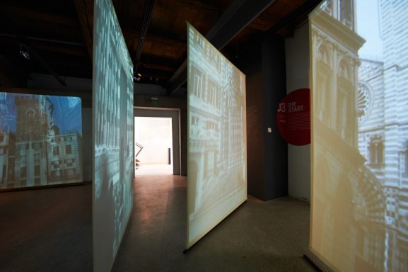 Ausstellungsraum mit Leinwänden, auf denen Filme von der Genueser Altstadt laufen