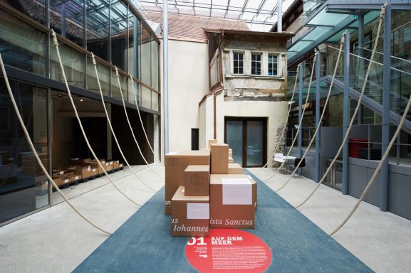 Foto des Innenhofs des Humpis-Quartiers, abstrahierte Darstellung eines Schiffsbauchs mit Hilfe von Tauen, Pappkisten mit lateinischen Namen